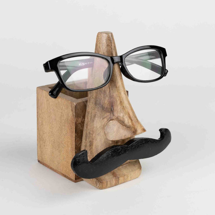 Handcrafted eyeglass and pencil holder made from sustainably sourced mango wood. Features a playful mustache face design in natural and black finishes. Measures 5.5" high with a 2.5" square base. Eco-friendly and easy to clean.