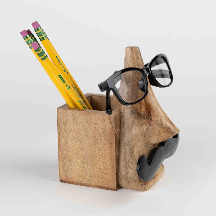 Handcrafted eyeglass and pencil holder made from sustainably sourced mango wood. Features a playful mustache face design in natural and black finishes. Measures 5.5" high with a 2.5" square base. Eco-friendly and easy to clean.
